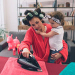 mother and baby together engaged in housework Ironing clothes . Housewife and kid doing homework. Woman with little child in the living room. Homemaker doing many tasks while looks after her infant.